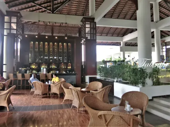 Spacious resort lounge with wicker chairs, wooden floors, and a bar area featuring glass cabinets.