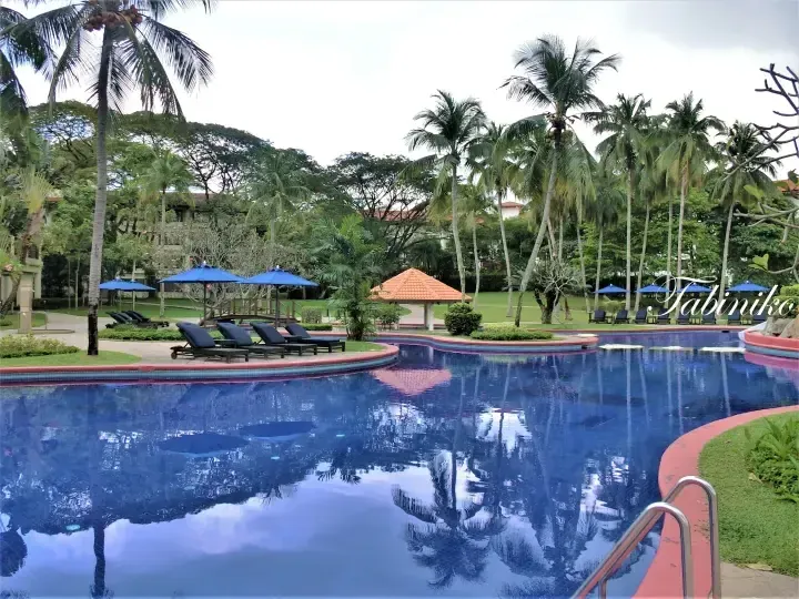 Resort swimming pool with blue umbrellas and palm trees in a lush tropical garden setting
