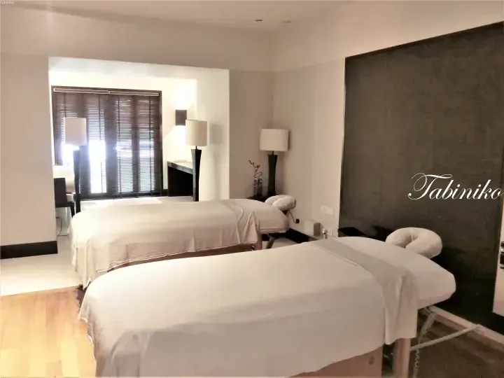 Two massage beds in a serene spa treatment room with wooden floors, lamps, and window blinds.