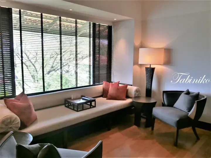 Modern hotel lounge with a long beige sofa, coral cushions, and large windows with dark blinds overlooking greenery.