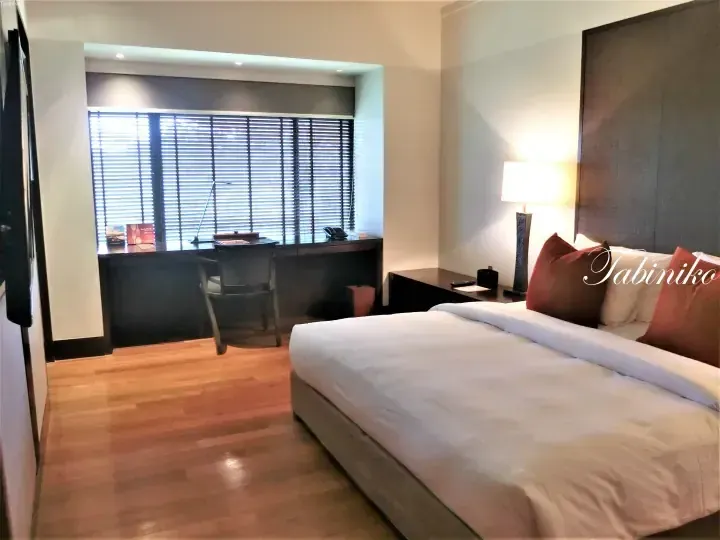 Modern hotel room with wooden floors, a large bed, and a desk by the window with blinds.