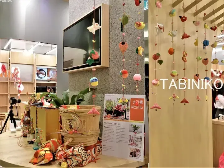 Colorful handmade Mizuhiki ornaments hanging at a TABINIKO display featuring a wooden barrel and event signage.
