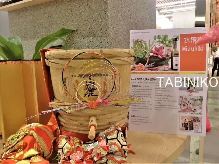 Traditional Japanese Mizuhiki decoration on a wooden bucket beside an informational display about Aya Nomura&rsquo;s Mizuhiki work.