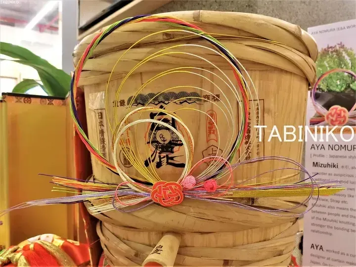 Colorful Mizuhiki cords tied in a decorative knot on a traditional wooden sake barrel at a TABINIKO display.