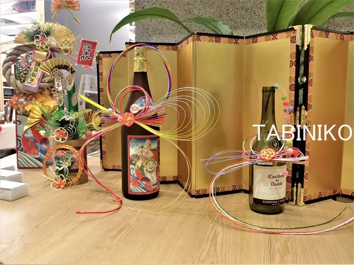 金色の屏風を背景に、祝祭的なリボンで飾られた日本酒とワインのボトルがテーブルに並んでいる。