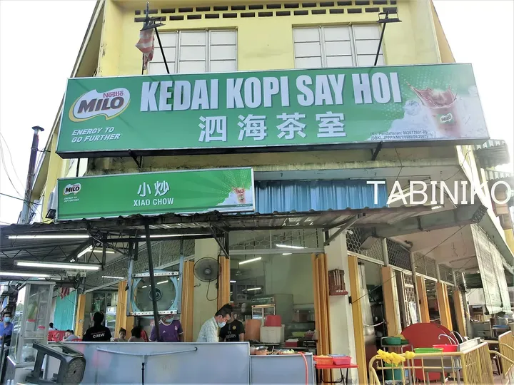 緑色の看板とMiloのロゴが目立つ「KEDAI KOPI SAY HOI」の外観。小炒（Xiao Chow）の屋台と客席が見える。