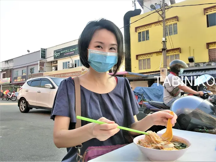 背景に黄色い建物とバイクが通るマレーシアの街で麺類を食べる女性。