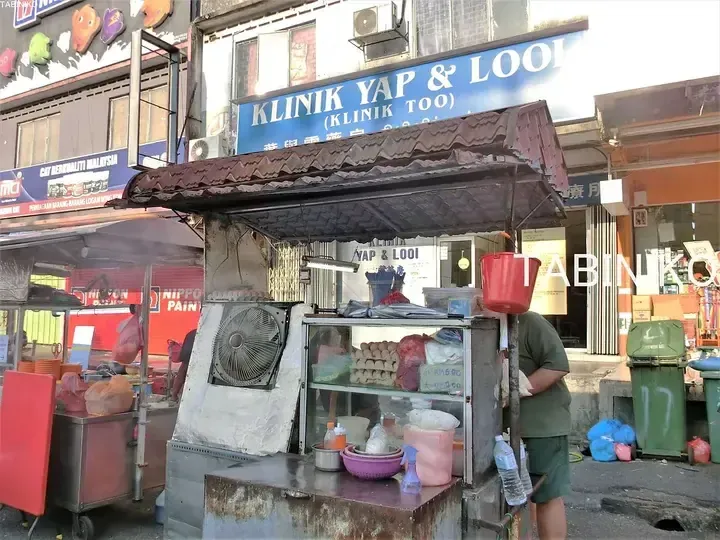 A street food stall in front of Klinik Yap & Looi in Malaysia, featuring a display of eggs and cooking equipment.