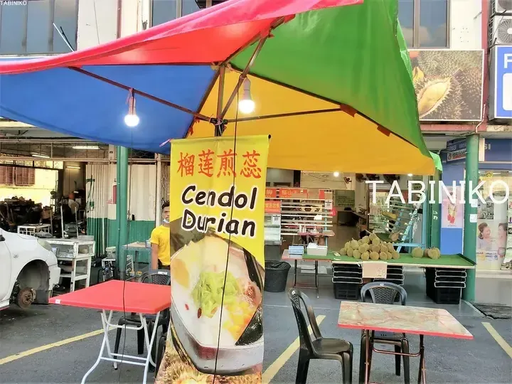 Colorful street stall selling Cendol Durian with fresh durians displayed on a table under a multi-colored canopy.