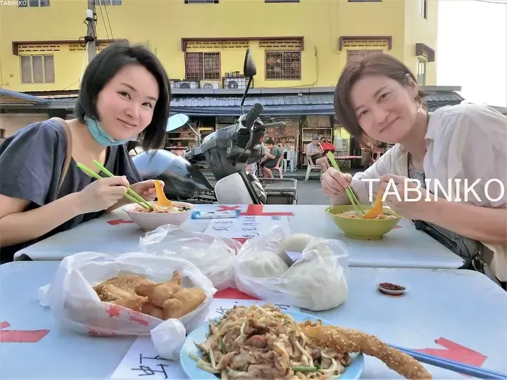 Two women enjoying local Malaysian street food including noodles and fried dough sticks at an outdoor table.