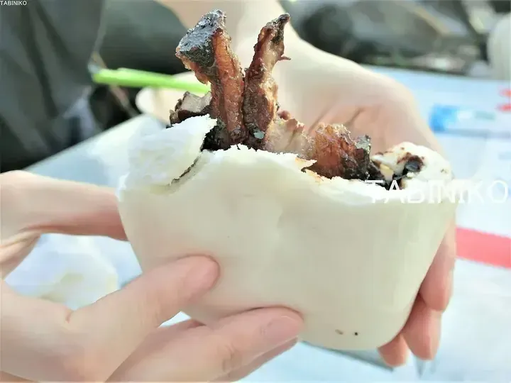 Hands holding a steamed bun stuffed with grilled meat, a popular Malaysian street food snack.