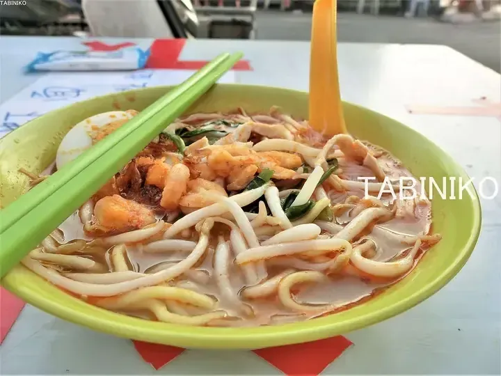 A bowl of laksa with prawns, egg, and bean sprouts in a yellow bowl with green chopsticks and a yellow spoon.