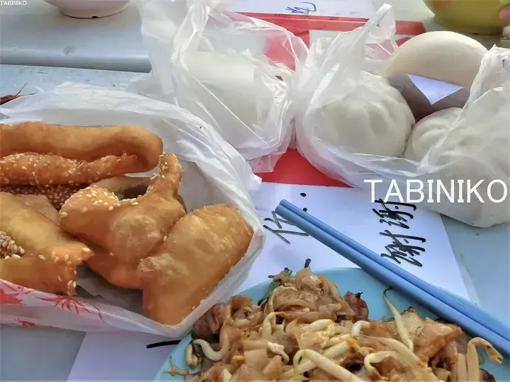 A table set with Chinese breakfast including fried dough sticks, steamed buns, and stir-fried rice noodles with chopsticks.