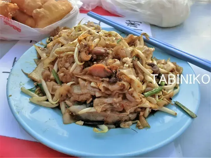 A plate of Malaysian char kway teow with bean sprouts, cockles, and tofu skin, served with blue chopsticks and a side of youtiao.