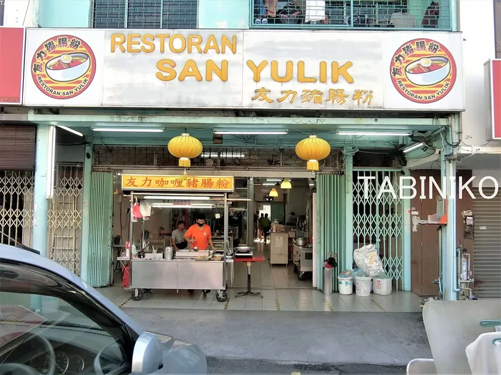 黄色い提灯が飾られたレストラン・サンユリク（Restoran San Yulik）の店舗外観と調理中のスタッフ。