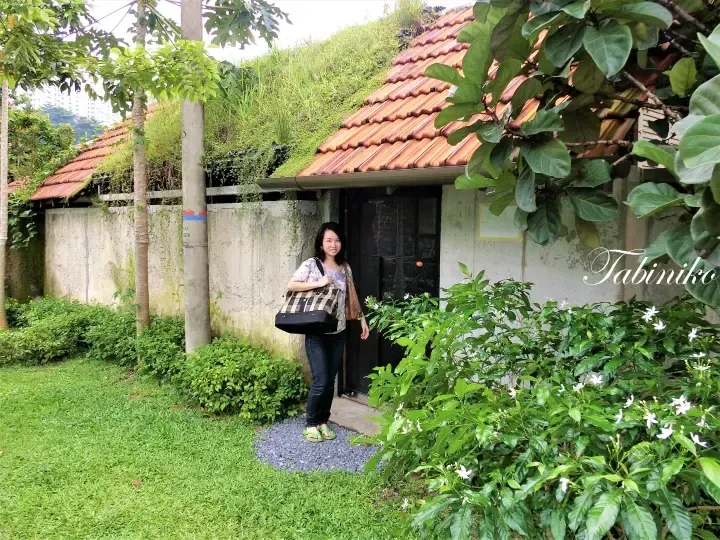 A woman stands in front of a green-roofed building with terracotta tiles in a lush garden setting.