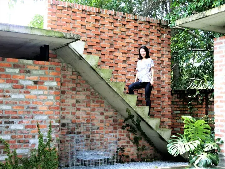 Woman posing on concrete stairs against a red brick wall at a modern property with lush greenery.