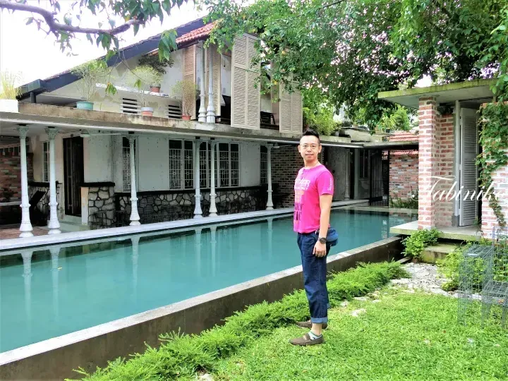 Man in pink shirt standing by a long pool at a heritage shophouse with white columns and brick details.
