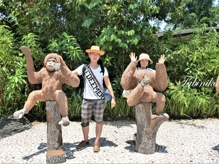A couple posing playfully with orangutan statues at a lush green park in Malaysia.