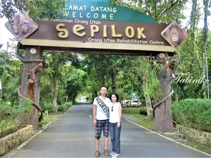A couple posing under the Sepilok Orang Utan Rehabilitation Centre entrance sign in Sandakan, Sabah, Malaysia.