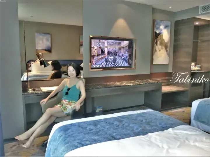 Woman relaxing in a modern hotel room with a TV showing H2O Pool Bar, marble desk, and blue bed accents.