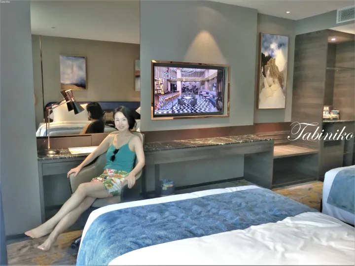 H2O Pool Barのテレビが映るモダンなホテル客室で、大理石のデスクと青いベッドアクセントが特徴の空間でリラックスする女性。