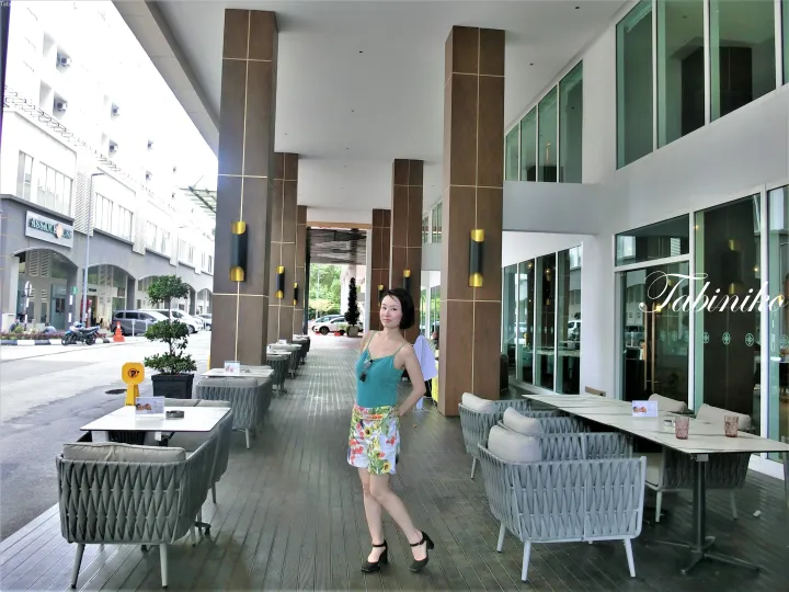 モダンなカフェのテラスで、大きな茶色の柱とグレーのウィッカー家具に囲まれた木製デッキに立つ女性。