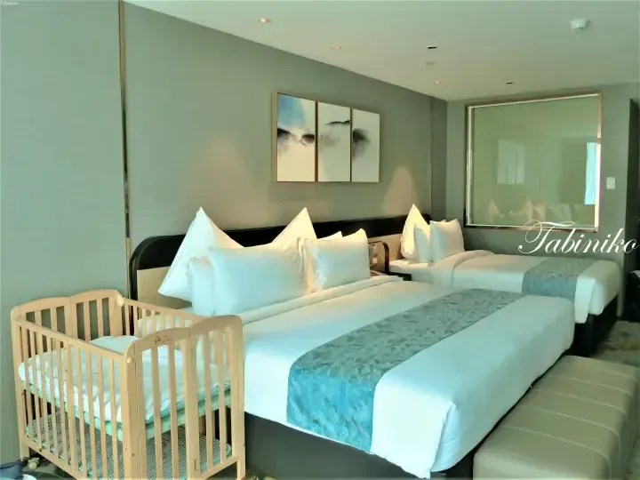 Modern hotel room with twin beds, blue accents, and a wooden crib for families.