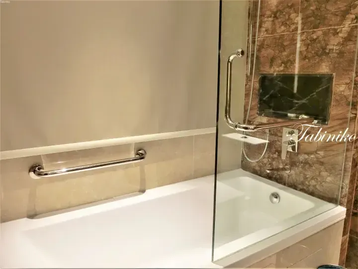 Modern hotel bathtub with beige marble walls, chrome grab bars, and a glass shower screen.