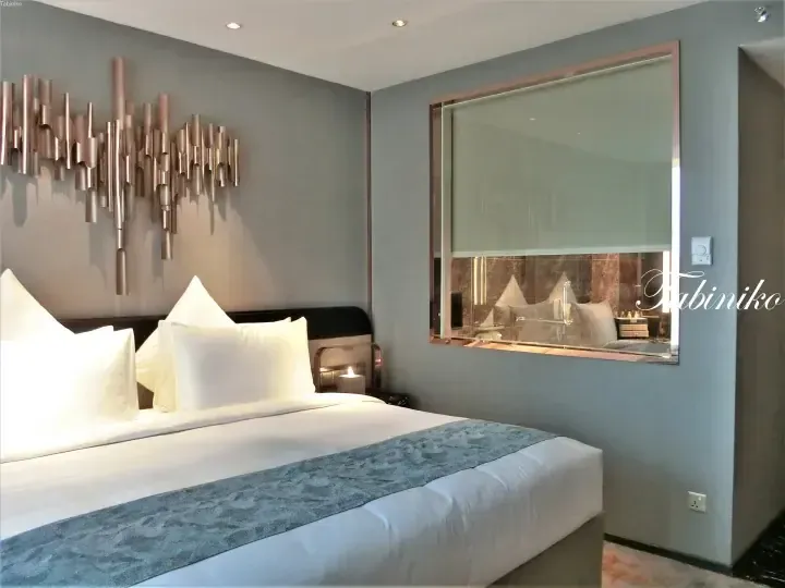 Modern hotel bedroom with a king bed, blue runner, and metallic wall art, featuring a glass partition to the bathroom.