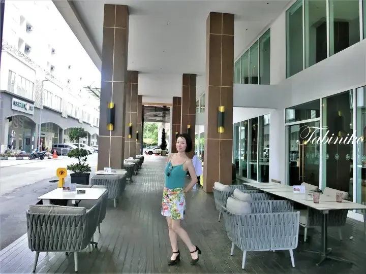 Woman posing on the wooden deck of a modern outdoor cafe terrace with grey wicker furniture and large brown pillars.