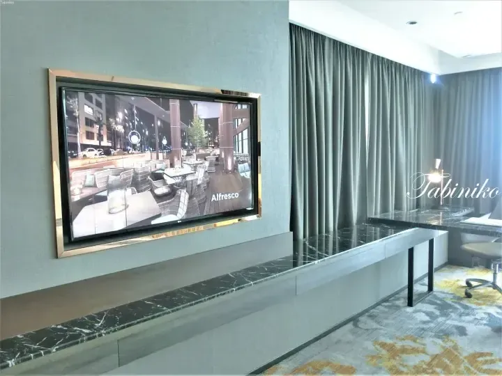 TV screen showing Alfresco restaurant interior in a modern hotel room with marble console and grey curtains.