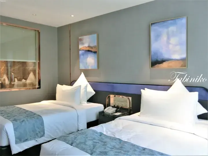 Modern hotel room with twin beds, blue artwork, and a glass bathroom partition.