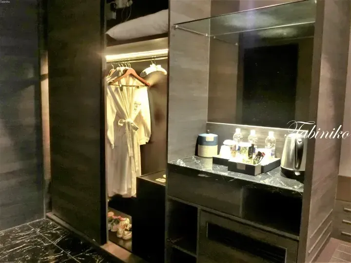 Modern hotel minibar area with kettle, water bottles, and white bathrobe hanging in open closet