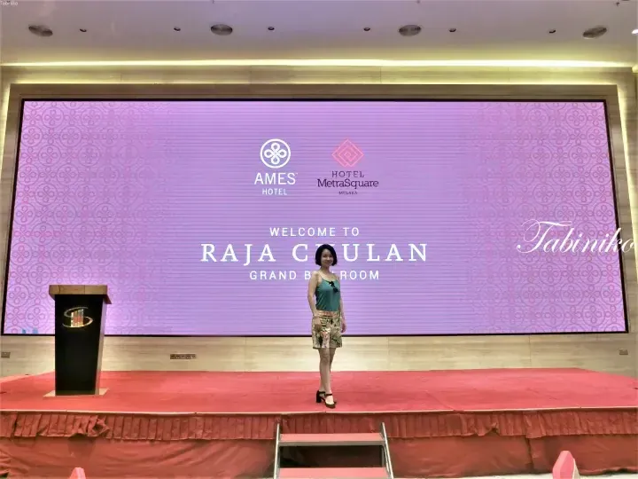 Woman posing on stage at Raja Chulan Grand Ballroom, Hotel MetraSquare Melaka, with AMES Hotel logo on screen.