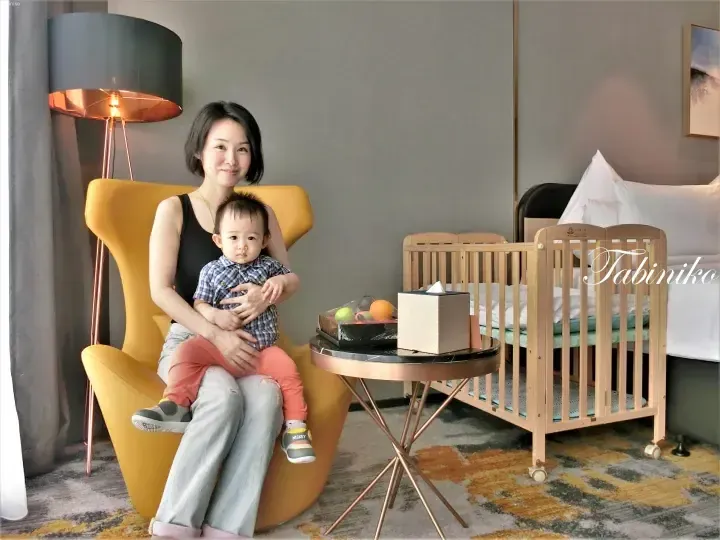 Mother and baby in a hotel room with a wooden crib, yellow armchair, and fruit basket.