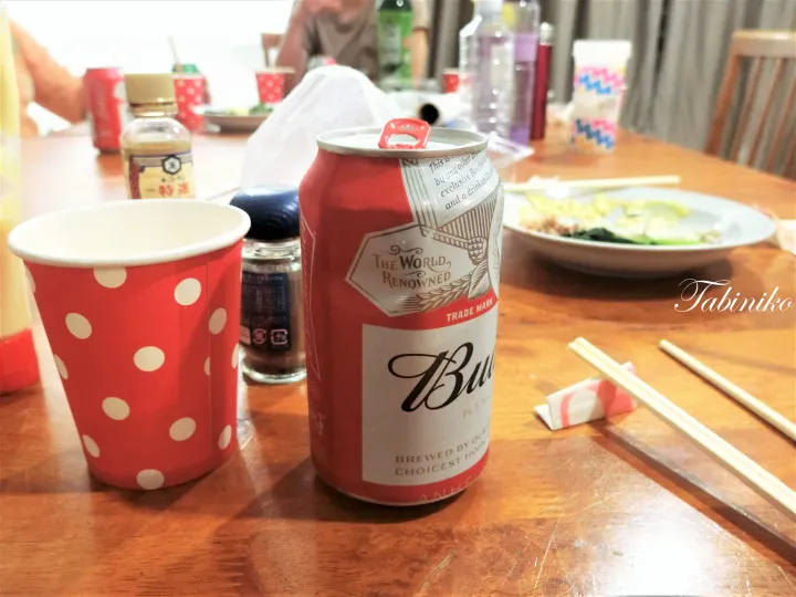 木製のダイニングテーブルに置かれた赤いバドワイザーの缶ビールと赤い水玉模様の紙コップ、箸、料理。