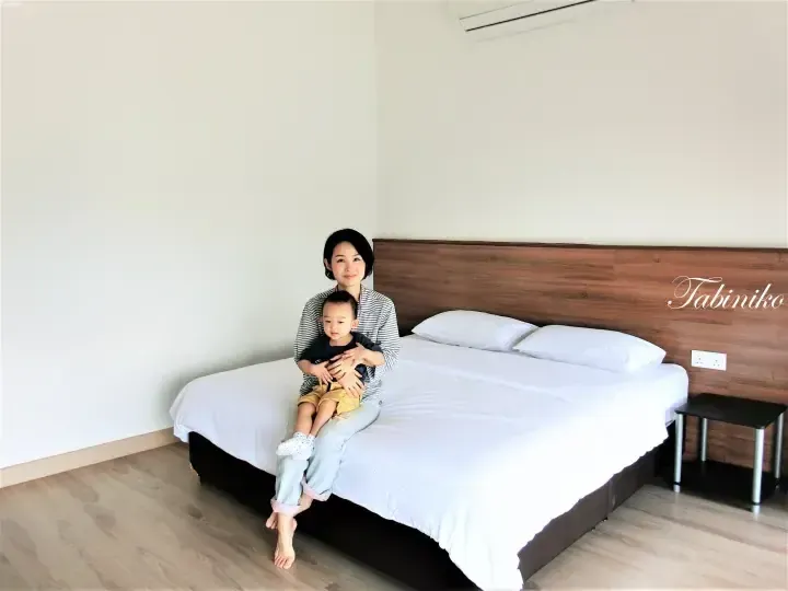A woman and child sitting on a hotel bed with white linens and a wooden headboard in a bright room.