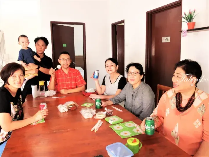 A group of adults and a child gather around a wooden table with snacks and Carlsberg drinks in a room with brown doors.