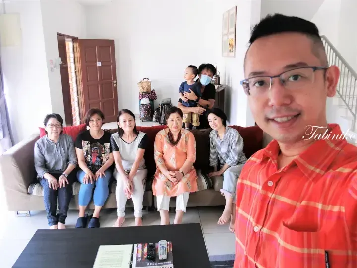 A family group selfie inside a bright living room with a sofa, luggage, and a staircase in the background.