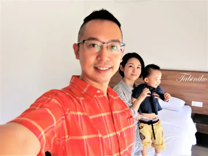 A family selfie in a hotel room with a smiling man in a red plaid shirt, a woman, and a toddler on a bed.