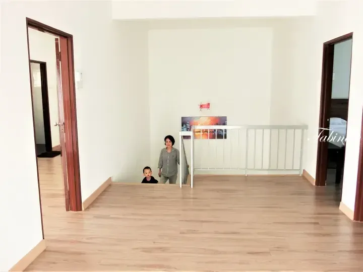 A woman and child stand at the top of a staircase in a bright, empty room with wooden flooring and white railings.
