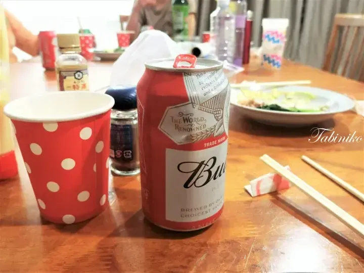 A red Budweiser beer can and a red polka-dot paper cup sit on a wooden dining table with chopsticks and plates of food.