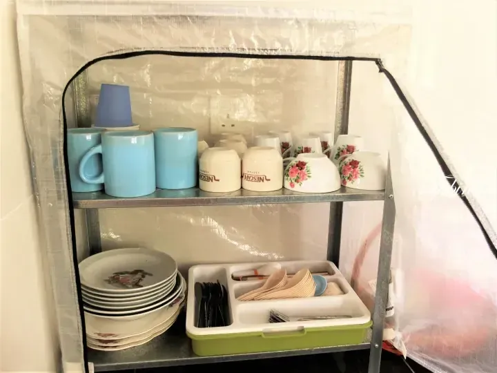 Organized dish rack with blue mugs, Nescafe cups, floral plates, and cutlery in a transparent cover.