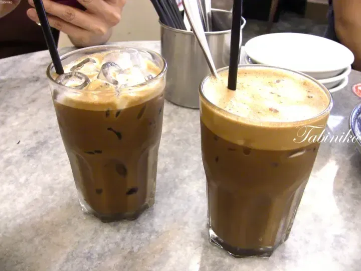 Two tall glasses of iced kopi with thick foam and black straws on a marble table at a Malaysian cafe.