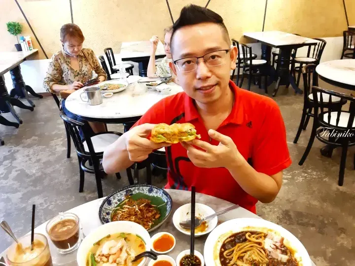 Man in red shirt holding a roti canai sandwich at a table with laksa and noodles in a Malaysian restaurant.