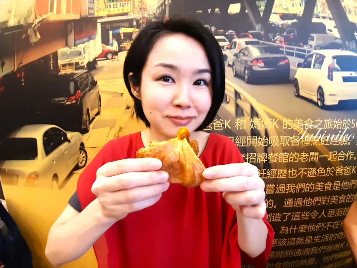 マレーシアの街並みと中国語のテキストが描かれた壁の前のモニュメントを前に、赤い服を着た女性が揚げ物を手にしている。
