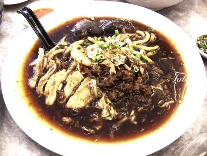 A bowl of dark noodle soup with chicken, minced meat, mushrooms, and spring onions.