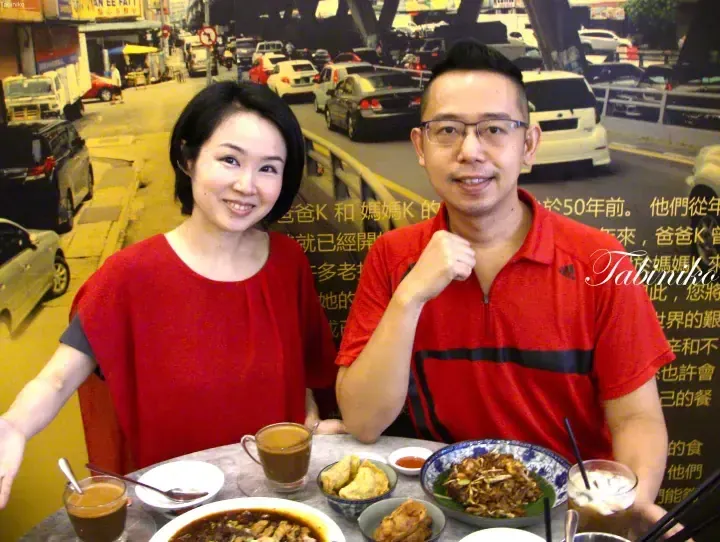 A couple smiles while enjoying a meal at a restaurant with a street scene backdrop featuring text about Baba Nyonya heritage.
