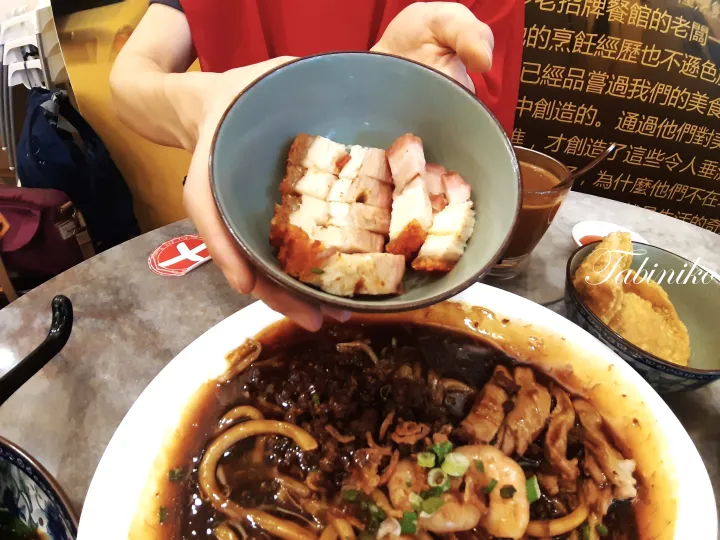 手が豚の角煮の皿を持ち、その下には海老や豚ひき肉が乗った大きな皿の麺料理が置かれている。